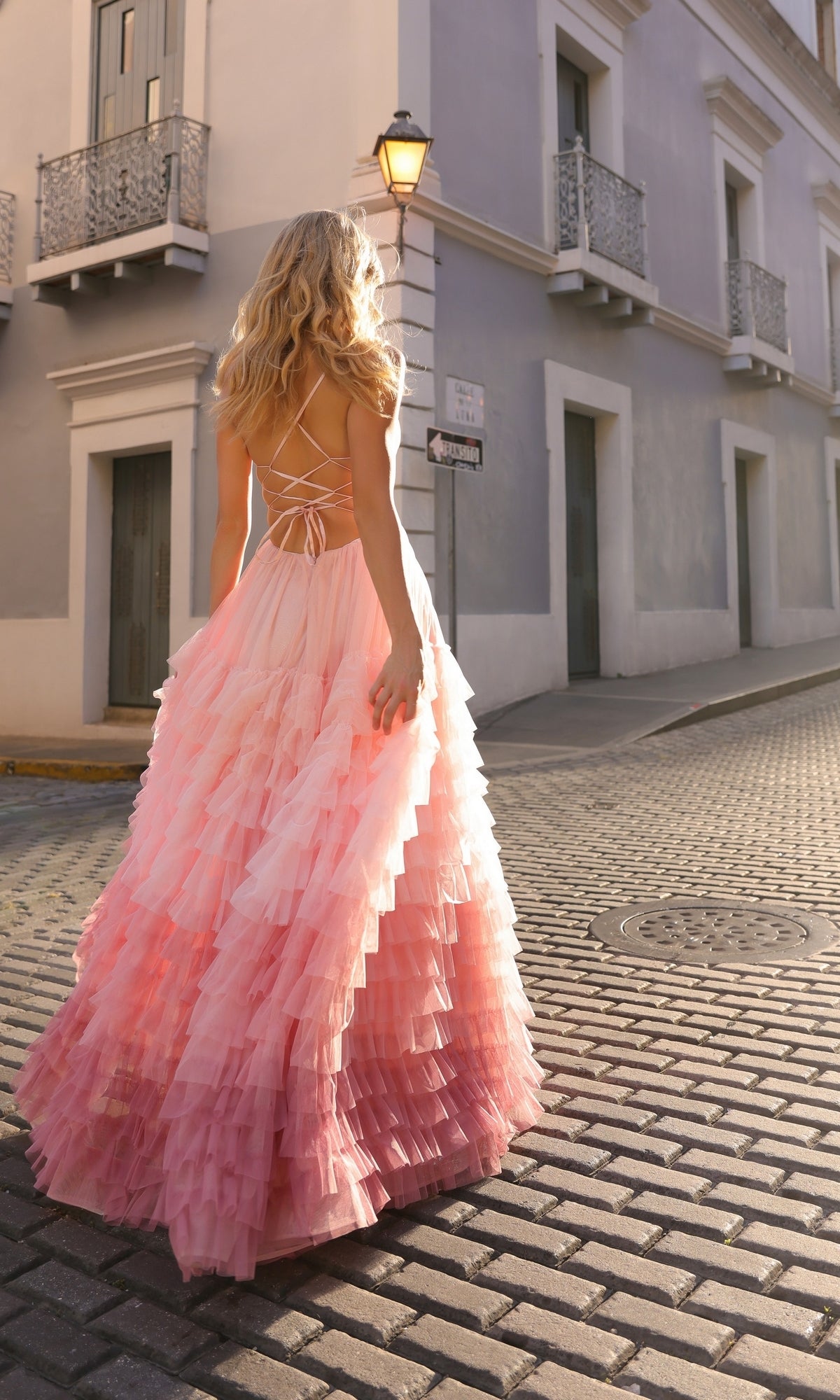 A woman with long blonde hair in the Nox Anabel Ruffled Ombre Prom Ball Gown C1420 walks alone on a cobblestone street beside a gray building.